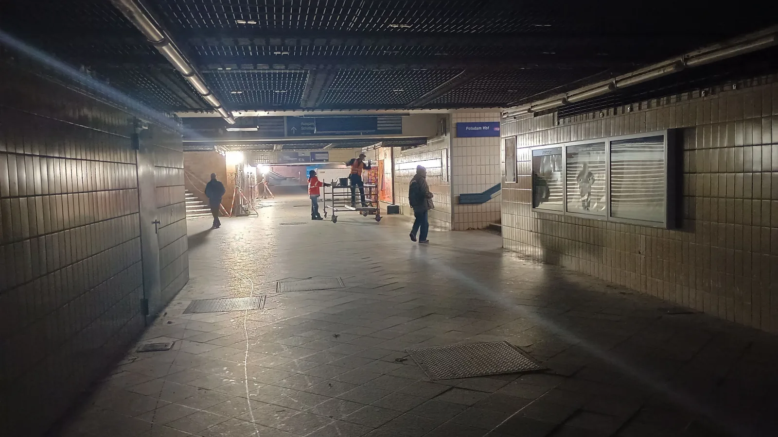 Eine dunkle Unterführung im Bahnhof Berlin-Wannsee, nur von Notbeleuchtung erhellt; Reisende gehen in der Ferne.