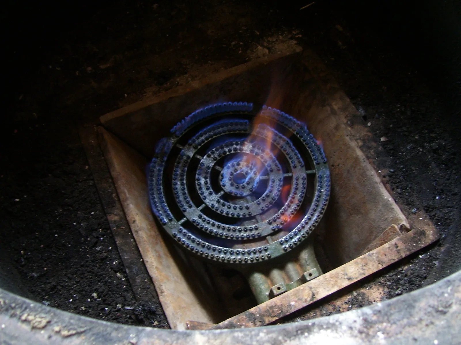 Ein runder gusseiserner Gasbrenner mit mehreren Düsenringen, eingelassen in eine gemauerte Kochstelle; aus den Düsen brennen kleine blaue Flammen, an einer Stelle züngelt eine orange Flamme hoch.