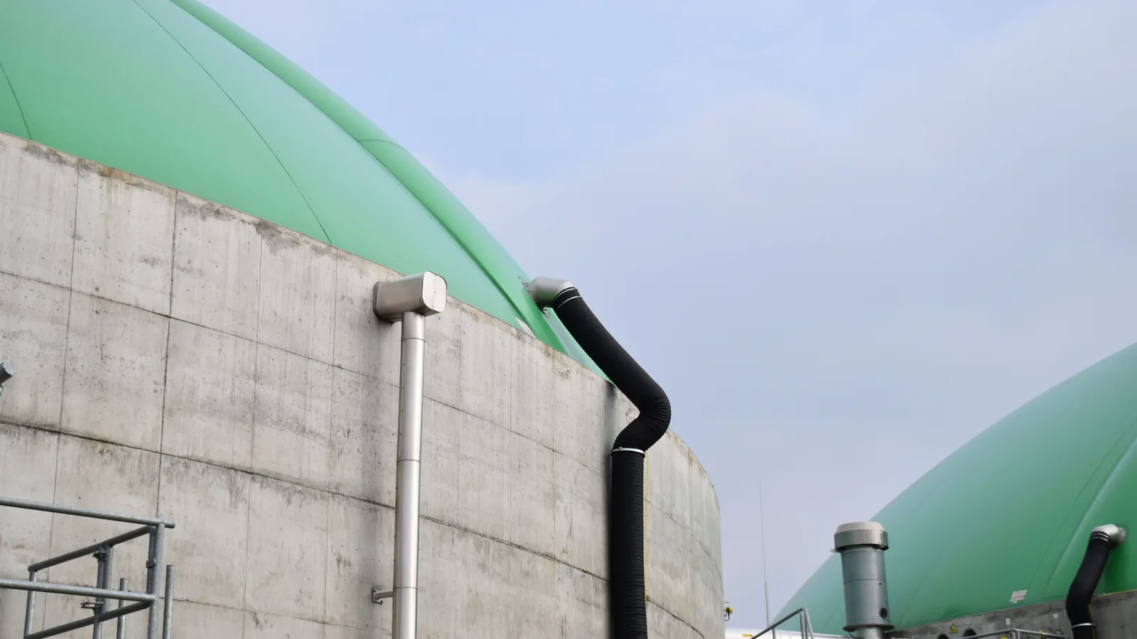 Runder Betonbehälter einer Biogasanlage mit grüner, gewölbter Folienhaube vor hellem Himmel; an der Wand laufen ein Metallrohr und ein schwarzer Luftschlauch nach oben, rechts dahinter ein zweiter Behälter.