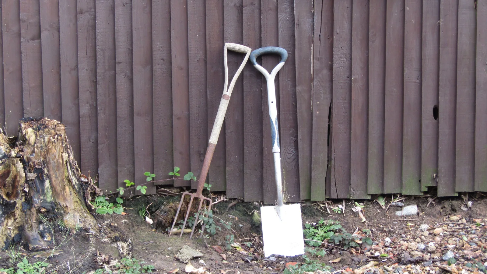 Ein Spaten und eine Grabegabel lehnen an einem dunklen Holzzaun; davor liegt frisch bearbeitete Gartenerde neben einem alten Baumstumpf.