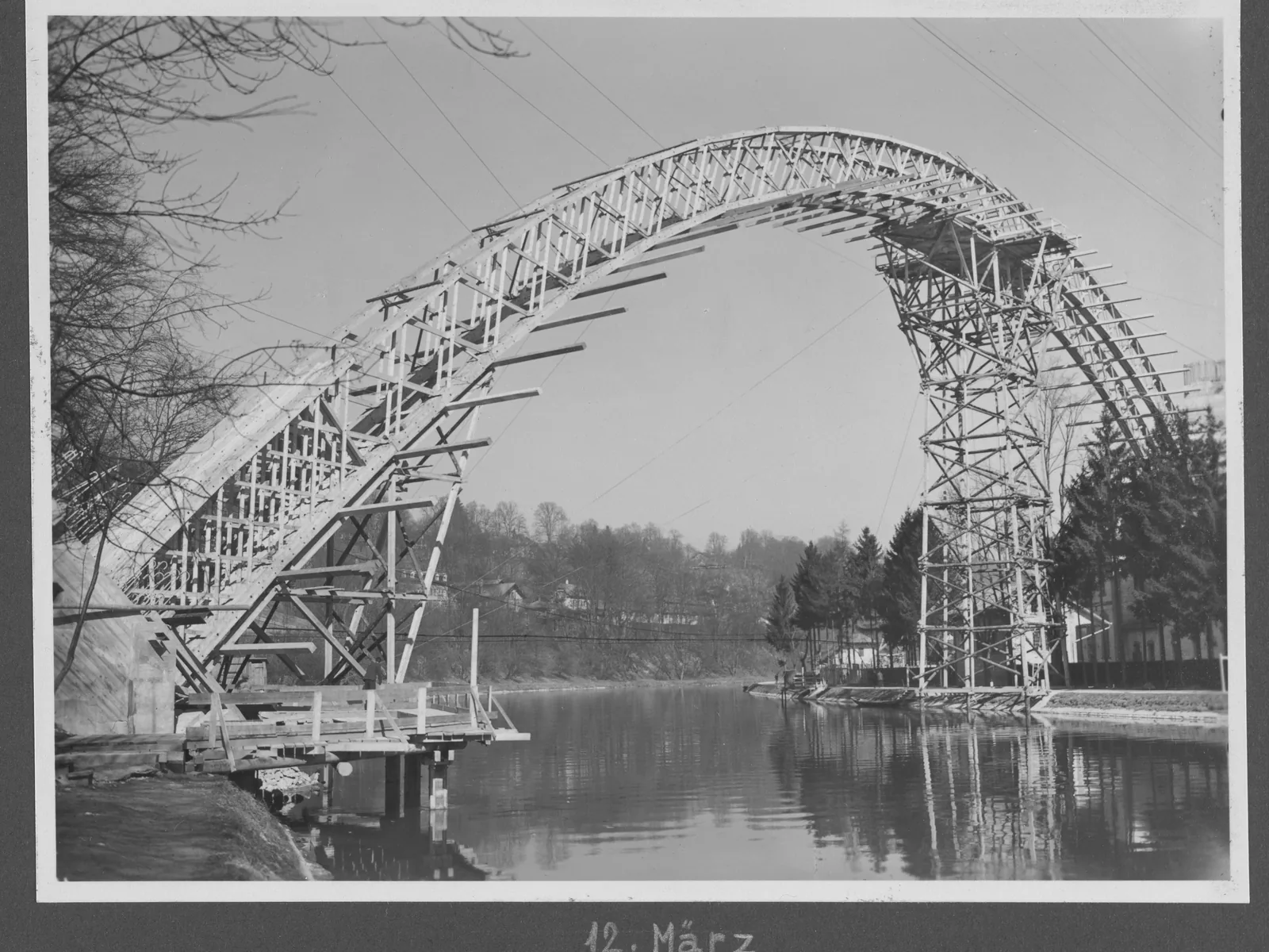 Historisches Schwarzweißfoto: Ein hölzernes Lehrgerüst in Bogenform spannt sich über einen Fluss, bevor der eigentliche Brückenbogen darauf gebaut wird.