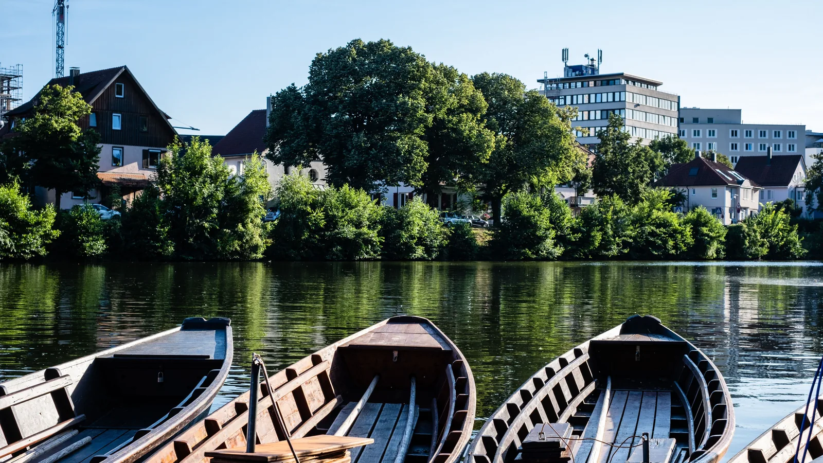 Mehrere leere, flache Holzkähne liegen nebeneinander an einem Steg auf ruhigem Flusswasser, im Hintergrund Bäume und Häuser am Ufer.