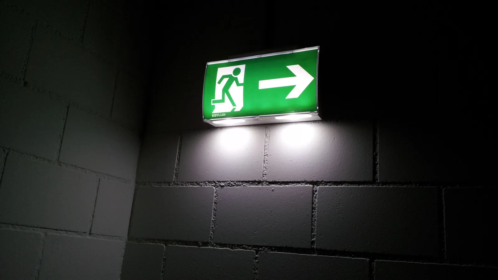 Ein beleuchtetes grünes Rettungszeichen mit laufender Person und Pfeil weist in einem dunklen Flur nach rechts.