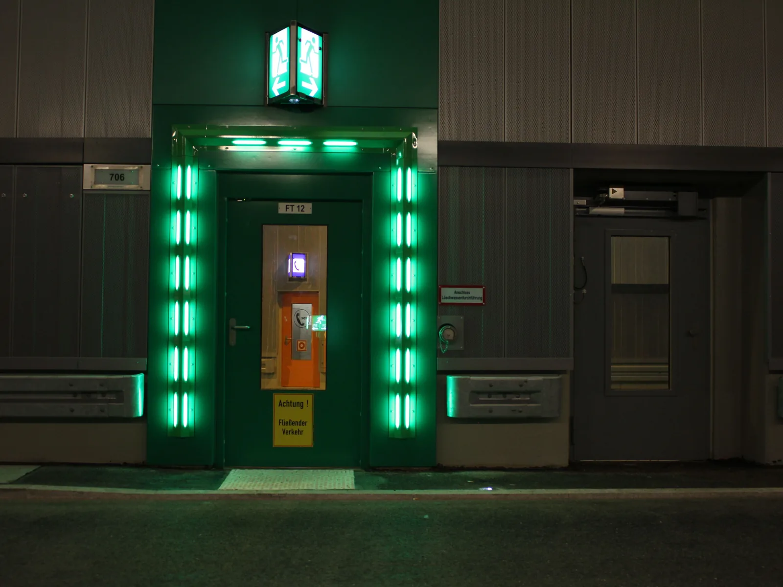 Grün beleuchtete Fluchttür in der Wand eines Straßentunnels, darüber ein Rettungszeichen.