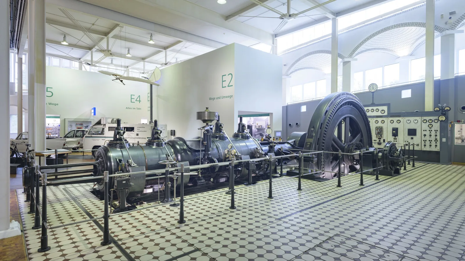 Eine große liegende Dampfmaschine mit mehreren Zylindern und einem schweren Schwungrad steht hinter einer Absperrung in einer hellen Museumshalle mit Fliesenboden.