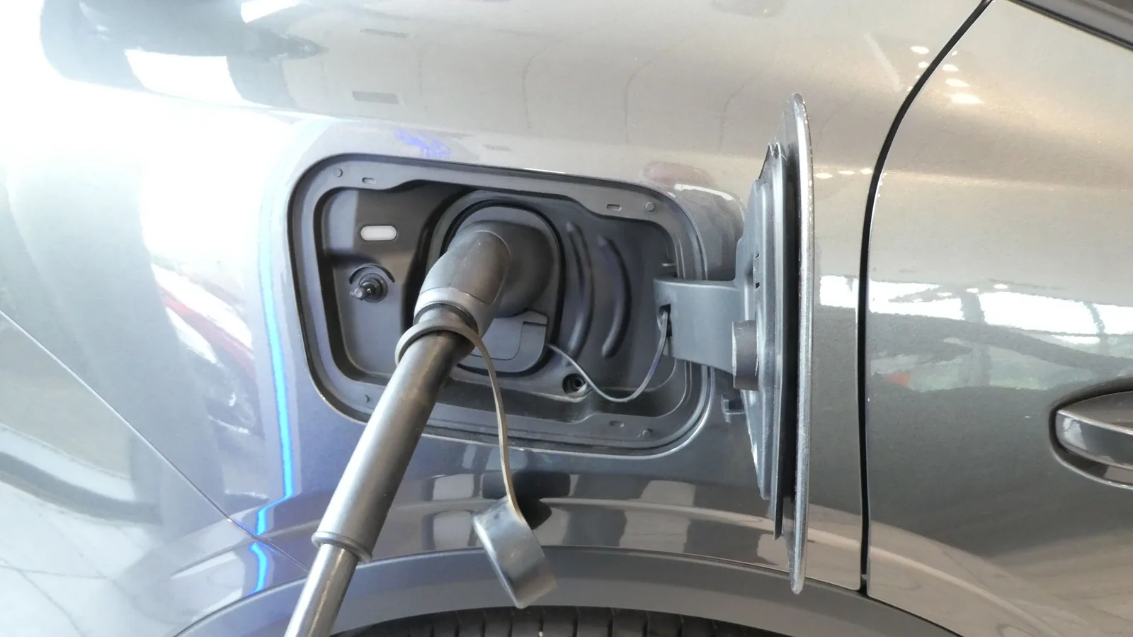 Nahaufnahme einer grauen Autoflanke mit geöffneter Ladeklappe, in der ein dunkler Ladestecker mit Kabel steckt.