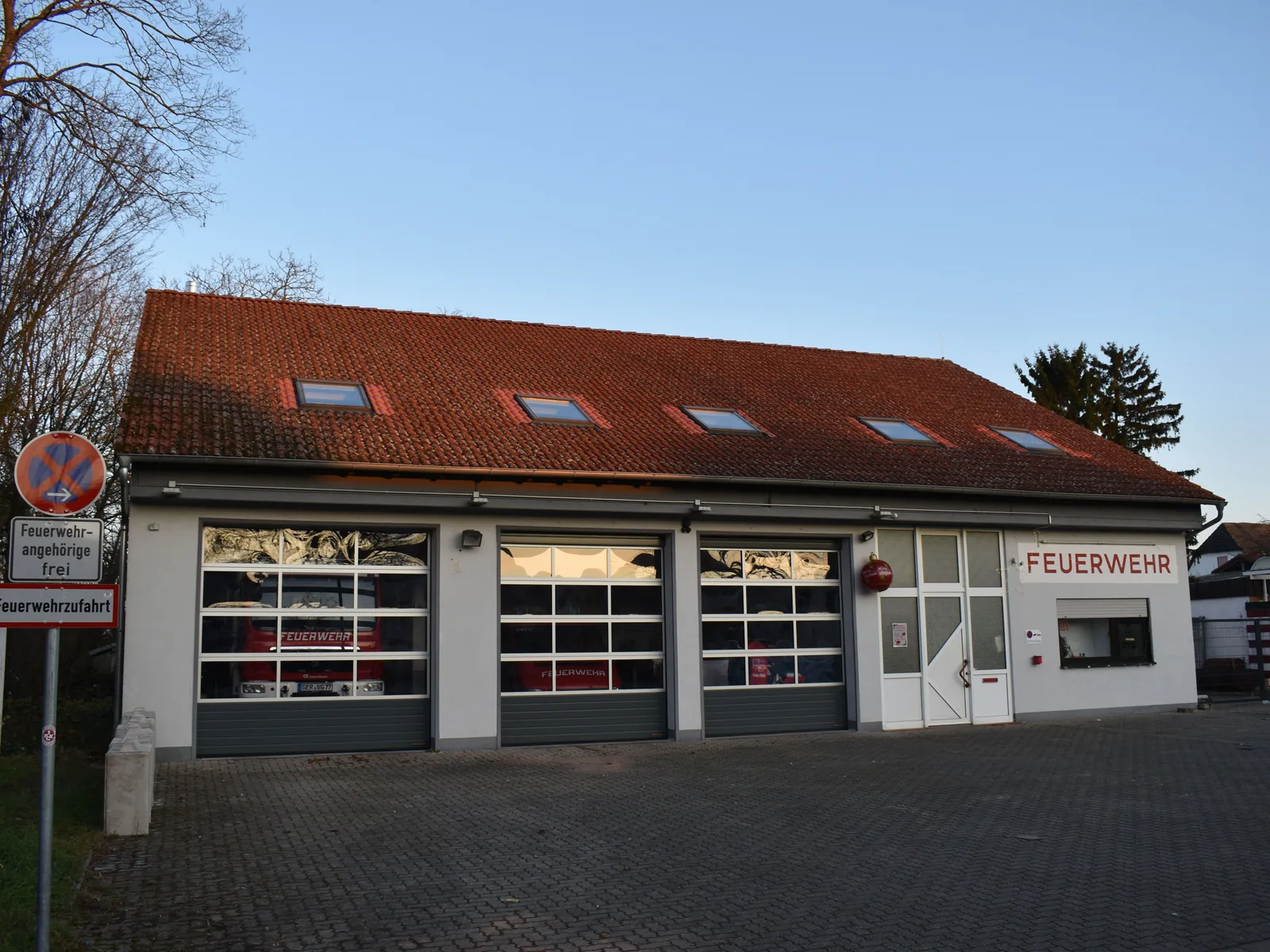 Gerätehaus einer Freiwilligen Feuerwehr in einem Dorf: ein Flachbau mit Ziegeldach, drei Rolltoren und der Aufschrift „Feuerwehr“, davor ein gepflasterter Hof und ein Schild „Feuerwehrzufahrt“.