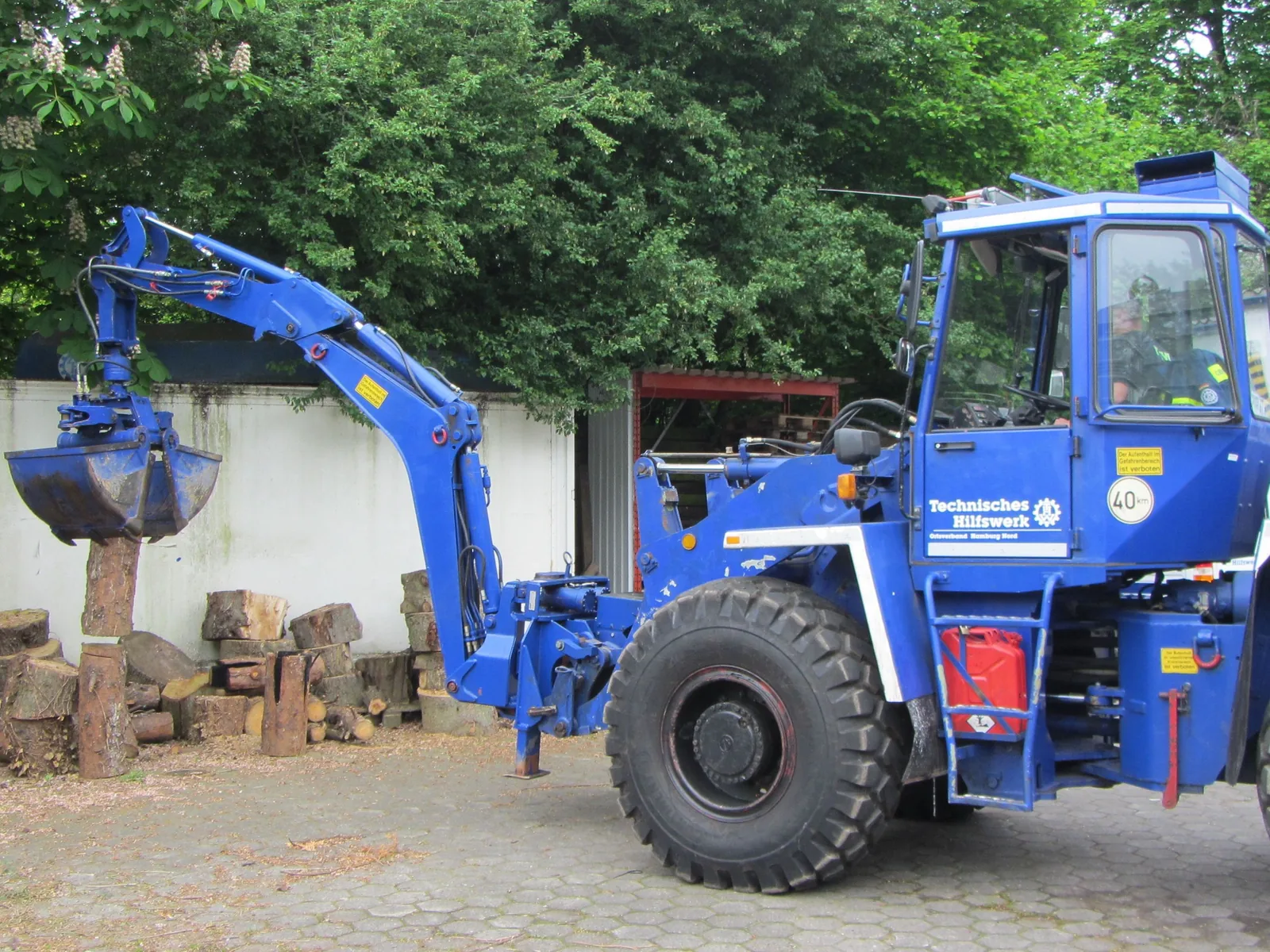 Ein blaues Bergungsräumgerät des Technischen Hilfswerks übt mit einem Zweischalengreifer das präzise Bewegen von Holzstücken.