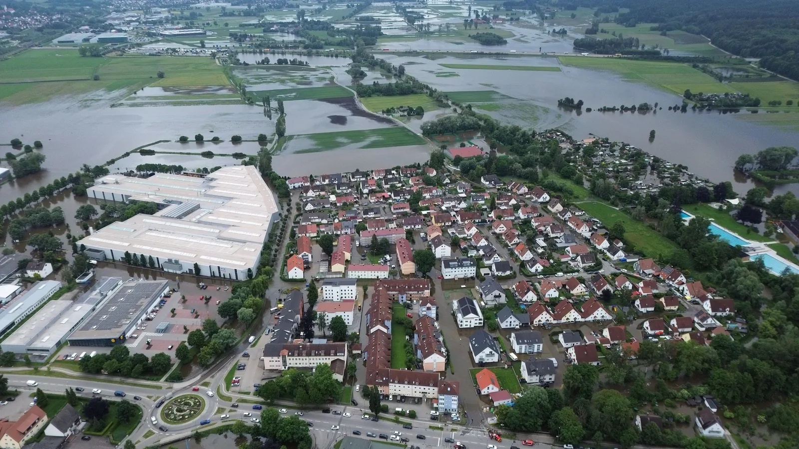 Luftbild von Burgau an der Mindel beim Hochwasser: Hinter dem Ort mit Einfamilienhäusern und Gewerbehallen stehen weite Wiesen und Felder großflächig unter Wasser, auch zwischen den Häusern steht Wasser.