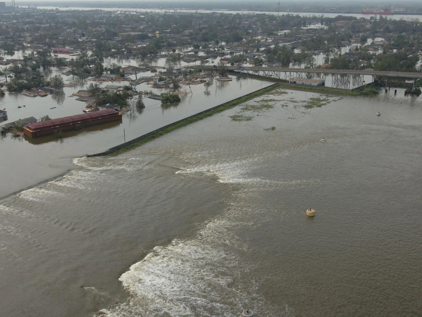 Luftbild einer gebrochenen Hochwasserschutzwand am Industrial Canal in New Orleans: Wasser strömt durch die Lücke, dahinter stehen Häuser bis zu den Dächern im Wasser.