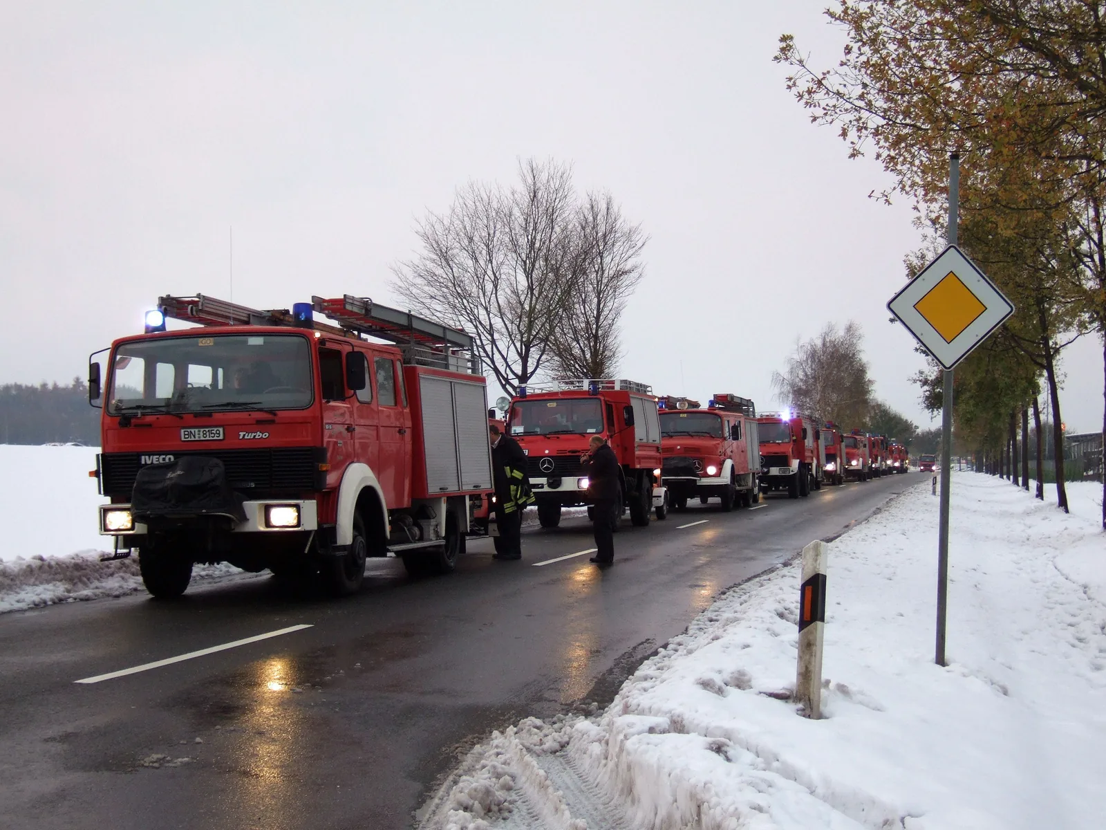 Eine lange Kolonne roter Feuerwehrfahrzeuge mit Blaulicht steht auf einer nassen Landstraße, am Rand liegt Schnee; zwei Einsatzkräfte stehen neben den Fahrzeugen.