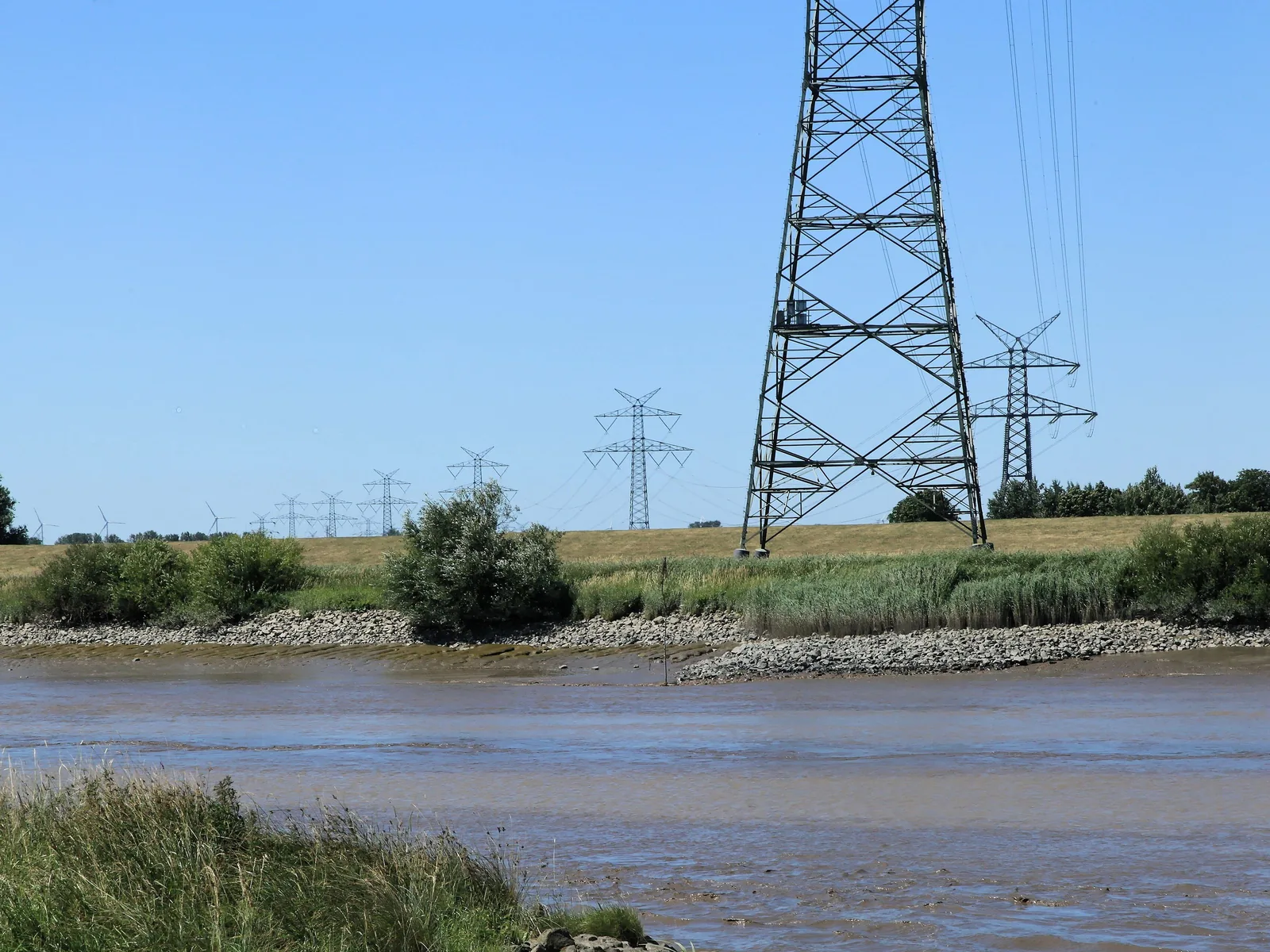Blick über die Ems bei Niedrigwasser auf einen hohen Stahlgittermast am anderen Ufer, dahinter weitere Masten einer Hochspannungsleitung, die sich über das flache Land zieht.
