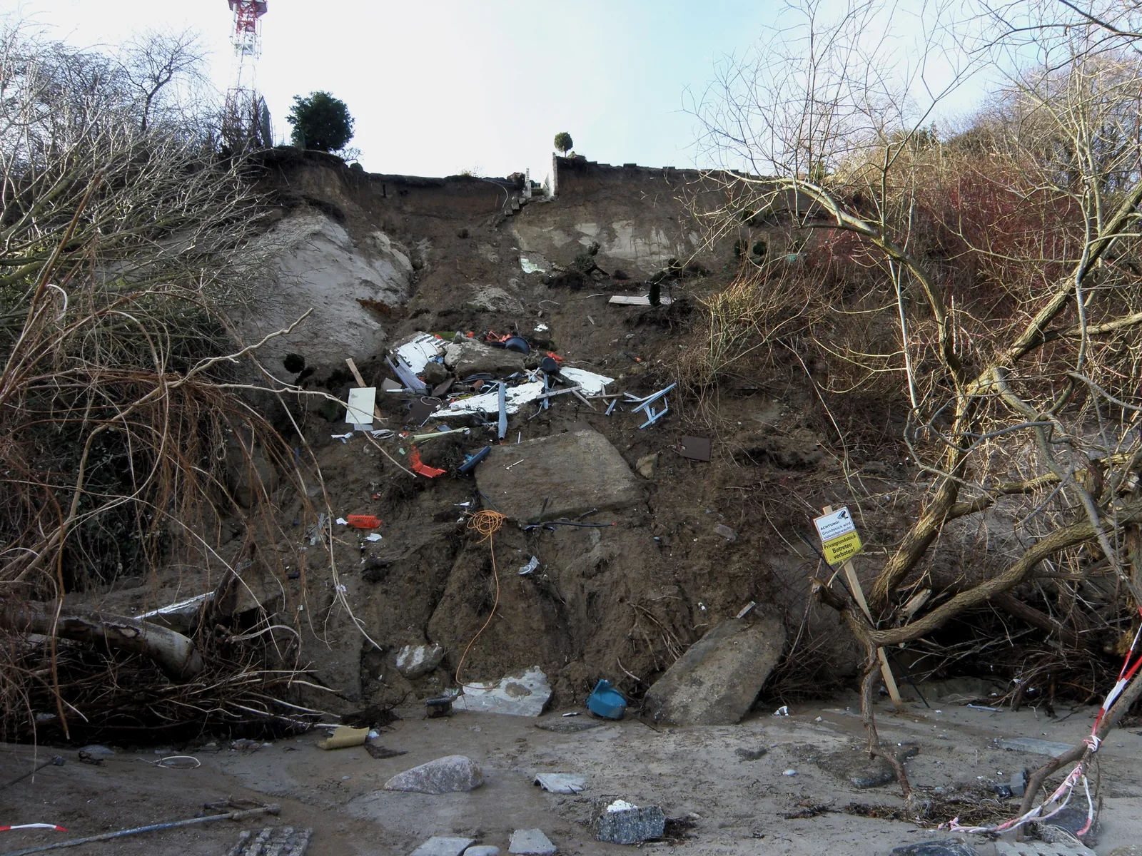 Ein Fußweg an der Kante eines Steilufers ist abgebrochen, Erde und Wurzeln sind abgerutscht, entwurzelte Bäume hängen zum Strand hinunter; im Hintergrund die Förde.
