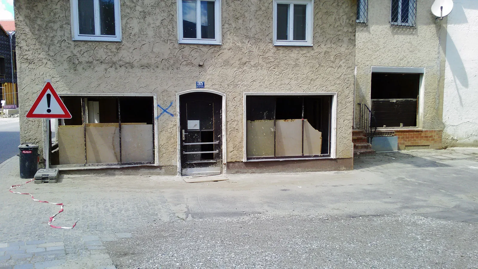 Hausfassade an der Innstraße in Simbach am Inn einige Wochen nach der Flut: Die Schaufenster im Erdgeschoss sind herausgebrochen und mit Platten verstellt, davor ein Warnschild und Absperrband, der Gehweg ist mit Schlamm und Kies bedeckt.