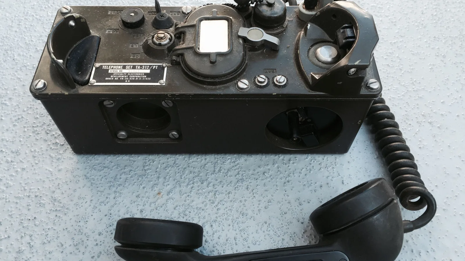 Ein geöffnetes US-Army-Feldtelefon TA-312 mit Hörer und Bedienelementen steht auf einer neutralen Unterlage.