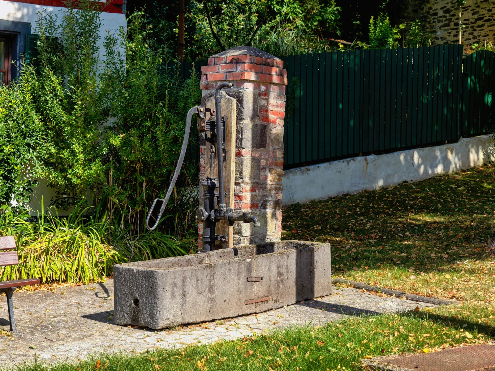 Gusseiserne Schwengelpumpe mit langem Hebel an einem gemauerten Pfeiler, davor ein langer Steintrog, im Hintergrund Garten und Zaun.