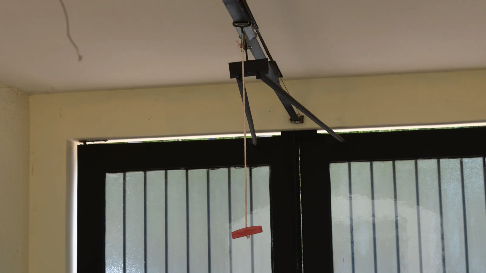 Laufschiene eines elektrischen Torantriebs an einer Garagendecke; von der Schiene hängt ein Seil mit rotem Griff zur Notentriegelung vor einem geschlossenen Tor.