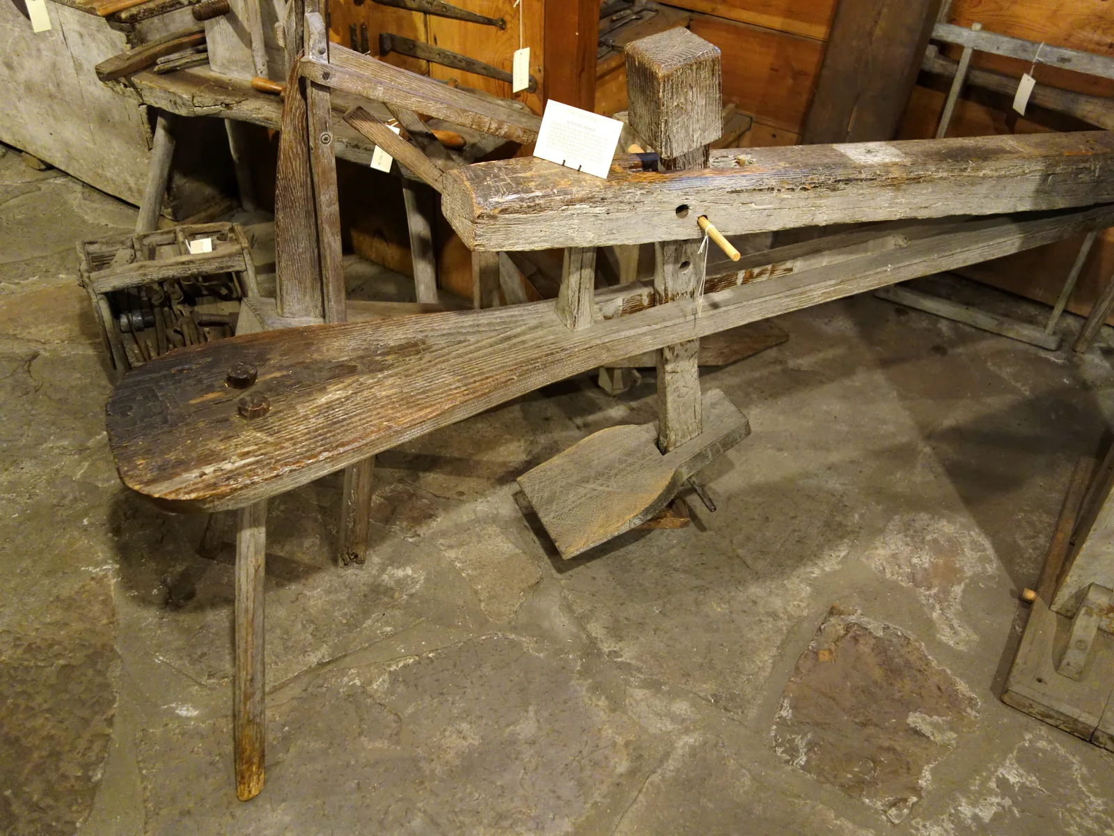 Eine alte hölzerne Schnitzbank im Museum: ein langer Sitzbalken auf Beinen, darüber ein Klemmbalken mit Kopfstück, der über ein Fußpedal niedergedrückt wird.