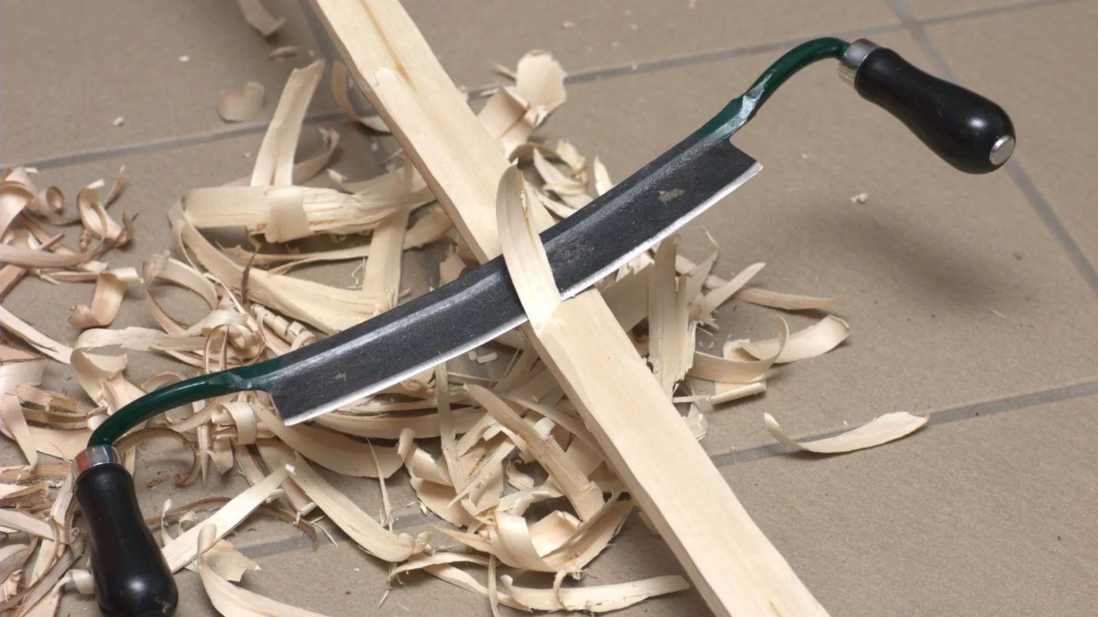 Ein Ziehmesser mit zwei Griffen liegt quer über einem hellen Holzstab, ringsum lange, gekringelte Späne.