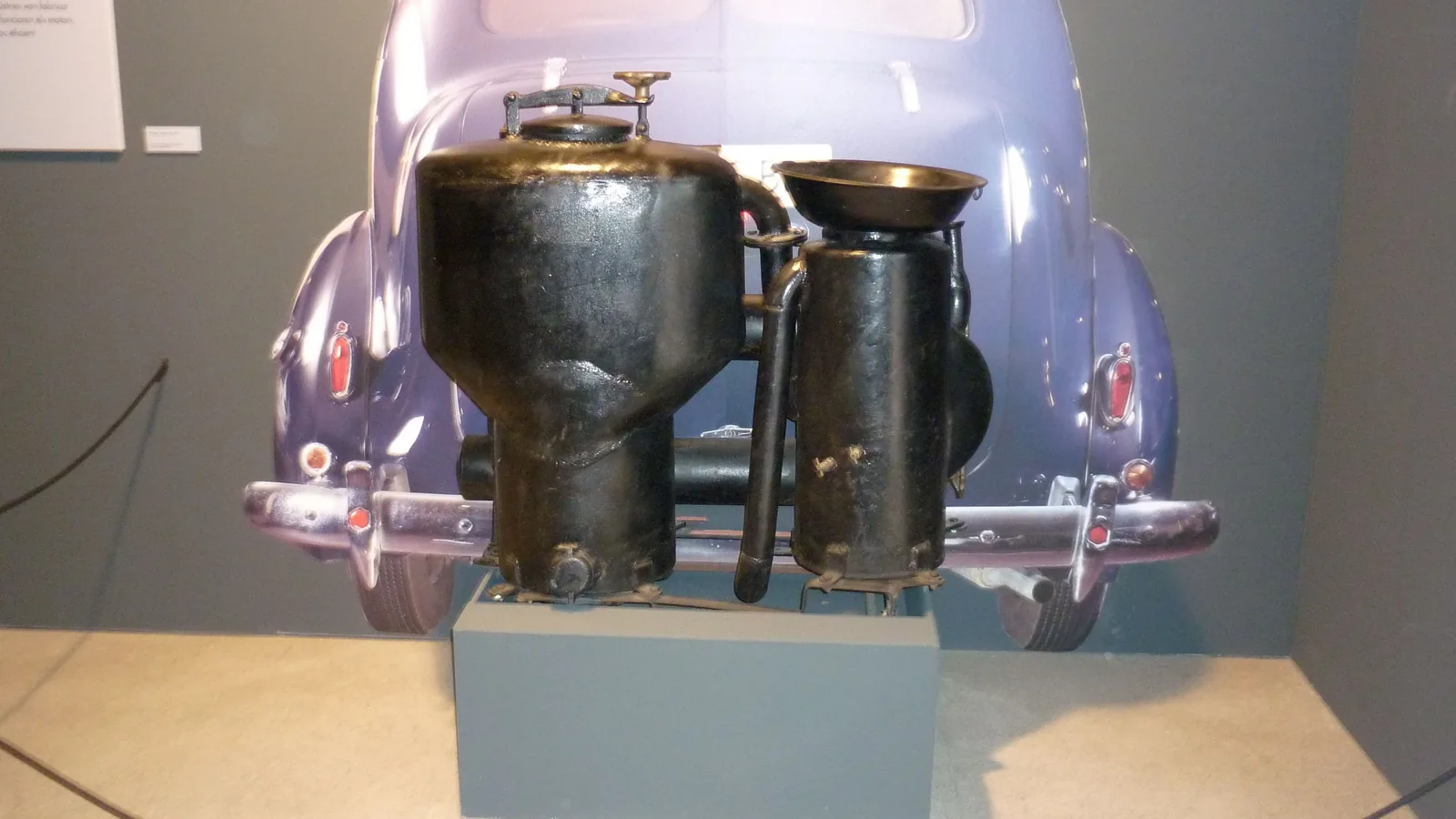 Museumsstück: zwei schwarze, rußige Metallbehälter mit Rohrverbindungen vor einer Fotowand mit dem Heck eines Personenwagens; links der bauchige Vergaser mit Deckel, rechts ein schlanker zweiter Behälter.