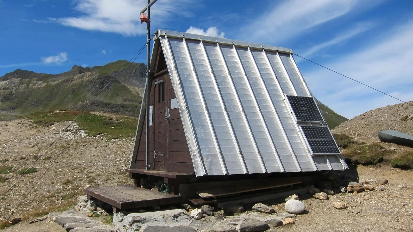Eine kleine Biwakhütte mit spitzem Blechdach steht auf einem steinigen Pass im Hochgebirge; auf der Dachfläche sind zwei kleine Solarmodule montiert.
