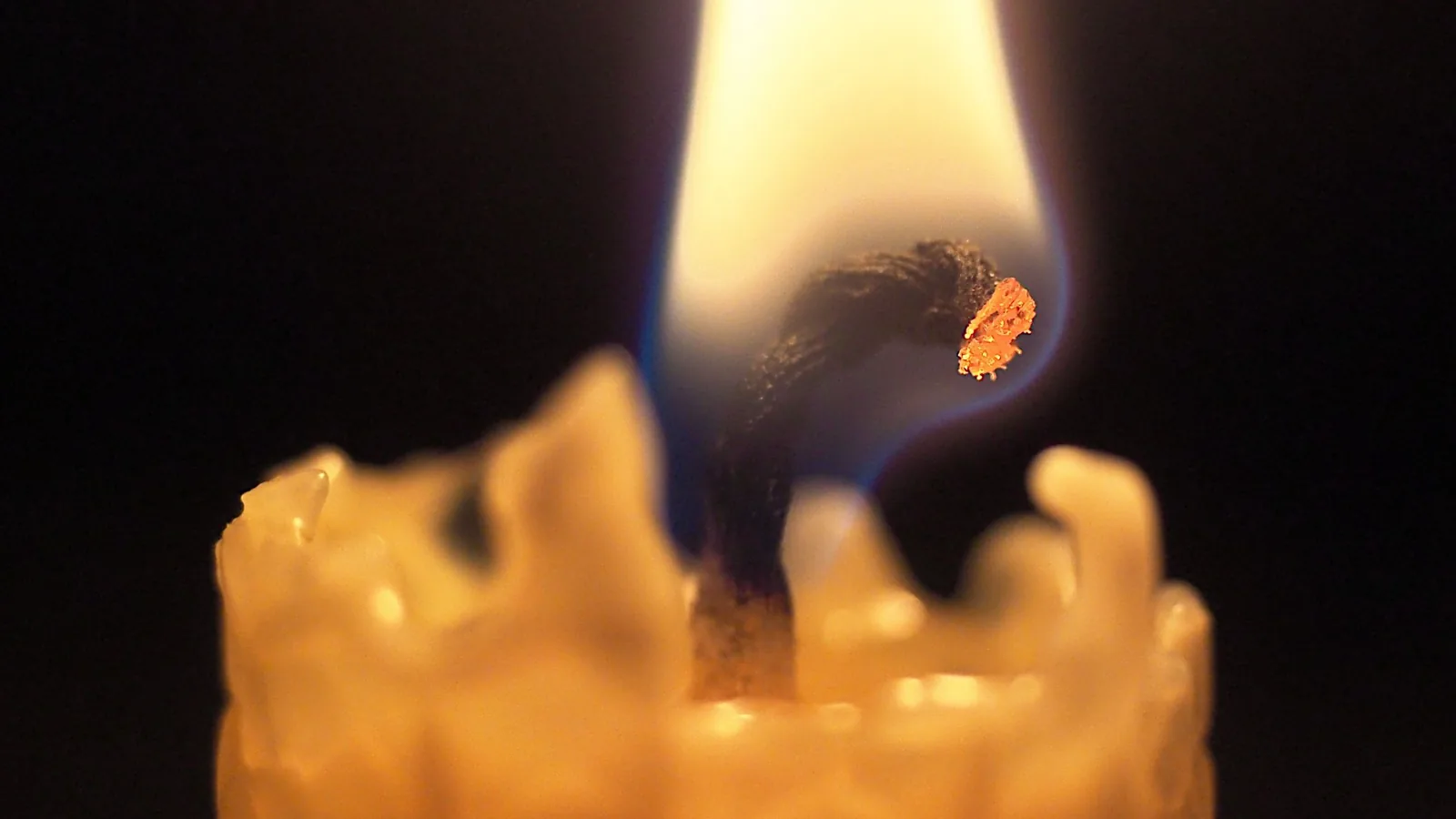Nahaufnahme einer brennenden Bienenwachskerze vor dunklem Hintergrund: Der gebogene Docht glüht an der Spitze, darum die gelbe Flamme mit bläulichem Saum, darunter der Rand aus geschmolzenem Wachs.