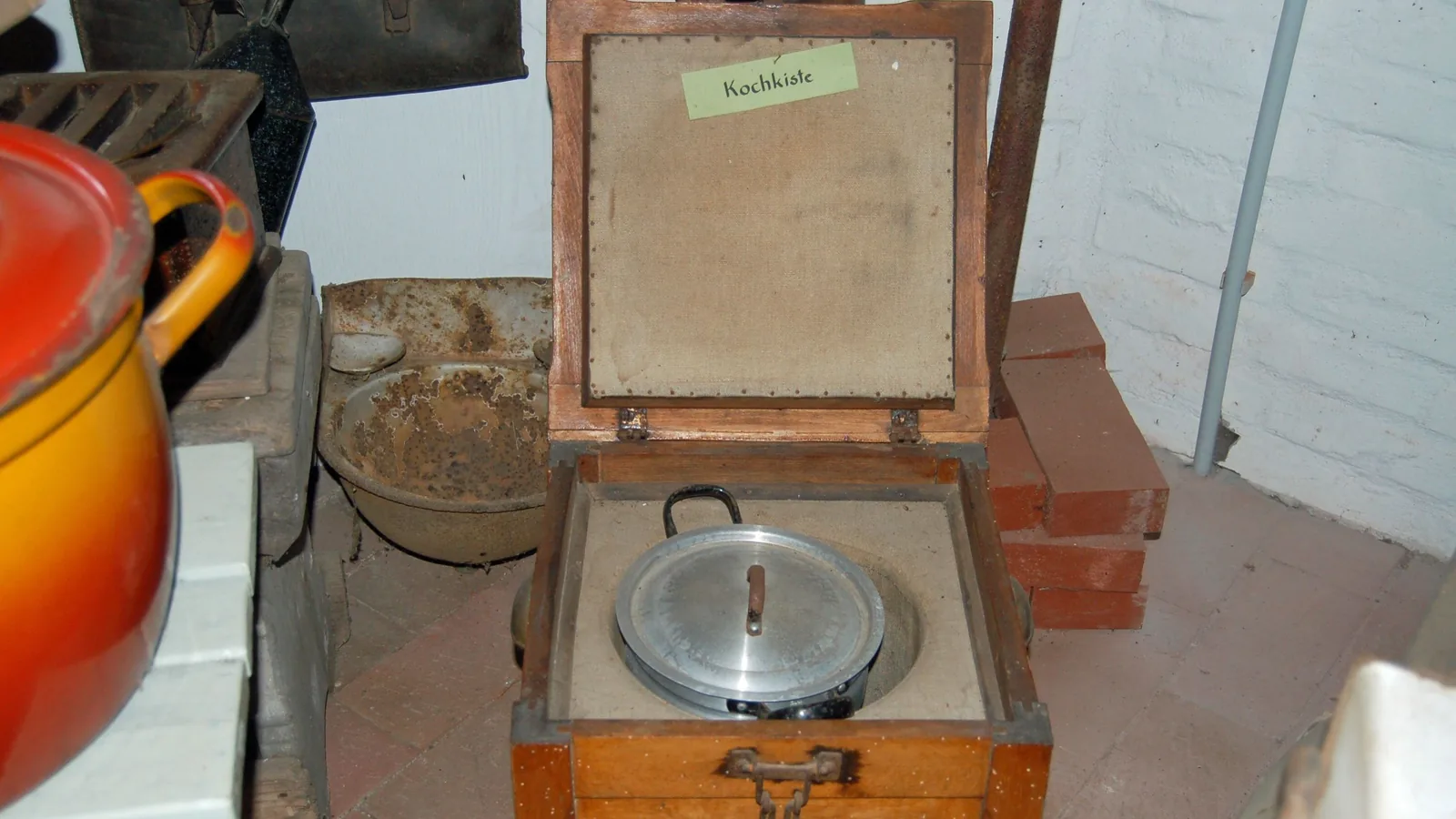 Eine historische Kochkiste aus Holz mit aufgeklapptem Deckel in einem Museum; in der gedämmten Mulde steht ein Aluminiumtopf mit Deckel.