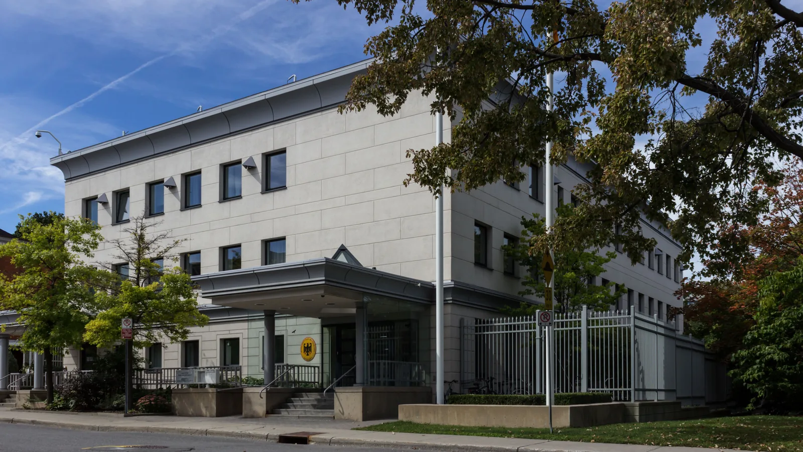 Das mehrstöckige Gebäude der deutschen Botschaft in Ottawa an einer Straßenecke, mit überdachtem Eingang, Zaun und Fahnenmast.