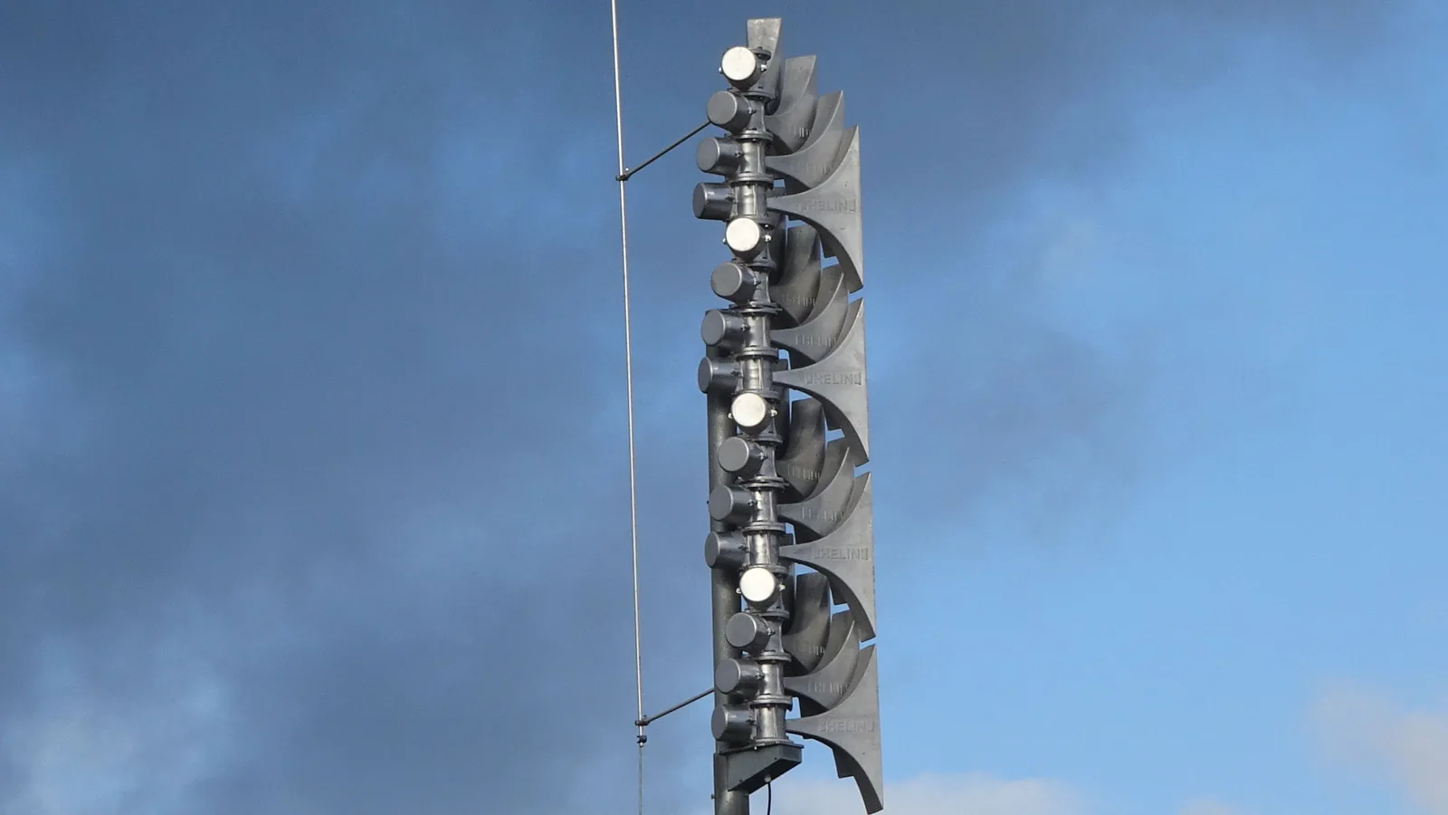 Eine elektronische Sirene mit übereinander angeordneten Schalltrichtern auf einem Mast über dem Schuldach in Altenbeken, vor blauem, teils bewölktem Himmel.