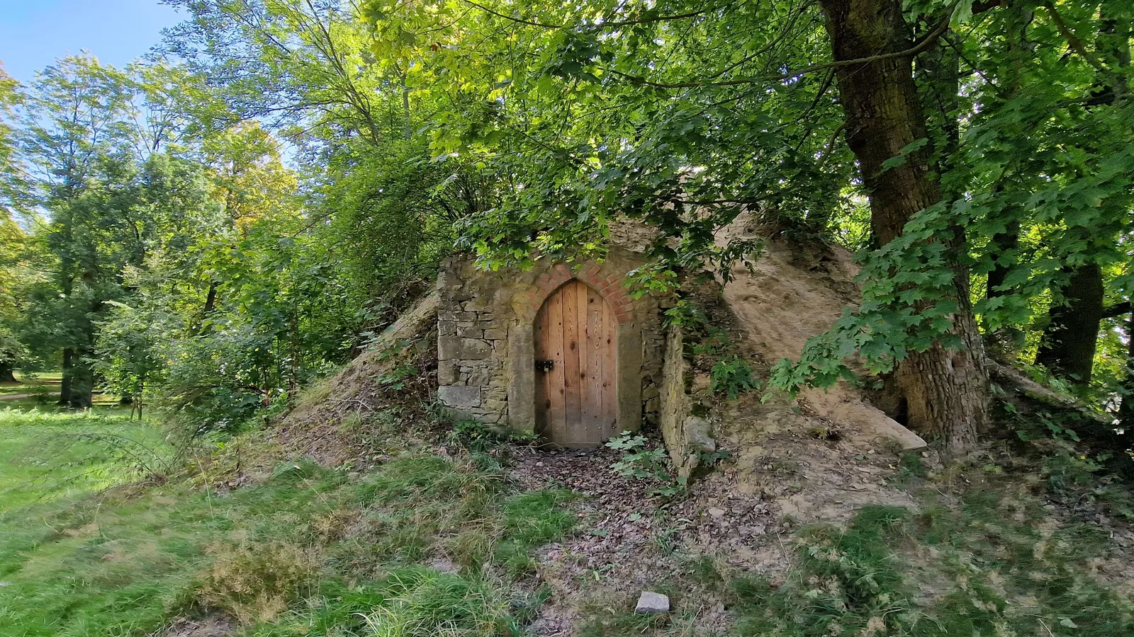 Ein historischer Eiskeller in einem Schlosspark: ein mit Erde und Gras überdeckter Hügel unter Bäumen, in dessen Front eine gemauerte Einfassung mit einer schmalen Holztür eingelassen ist.