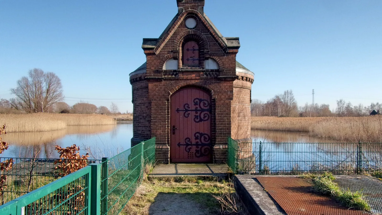 Ein kleines Backsteinhaus mit roter Tür steht auf einem Damm zwischen zwei ehemaligen Filterbecken, in denen Wasser und Schilf stehen; im Vordergrund ein grüner Zaun.