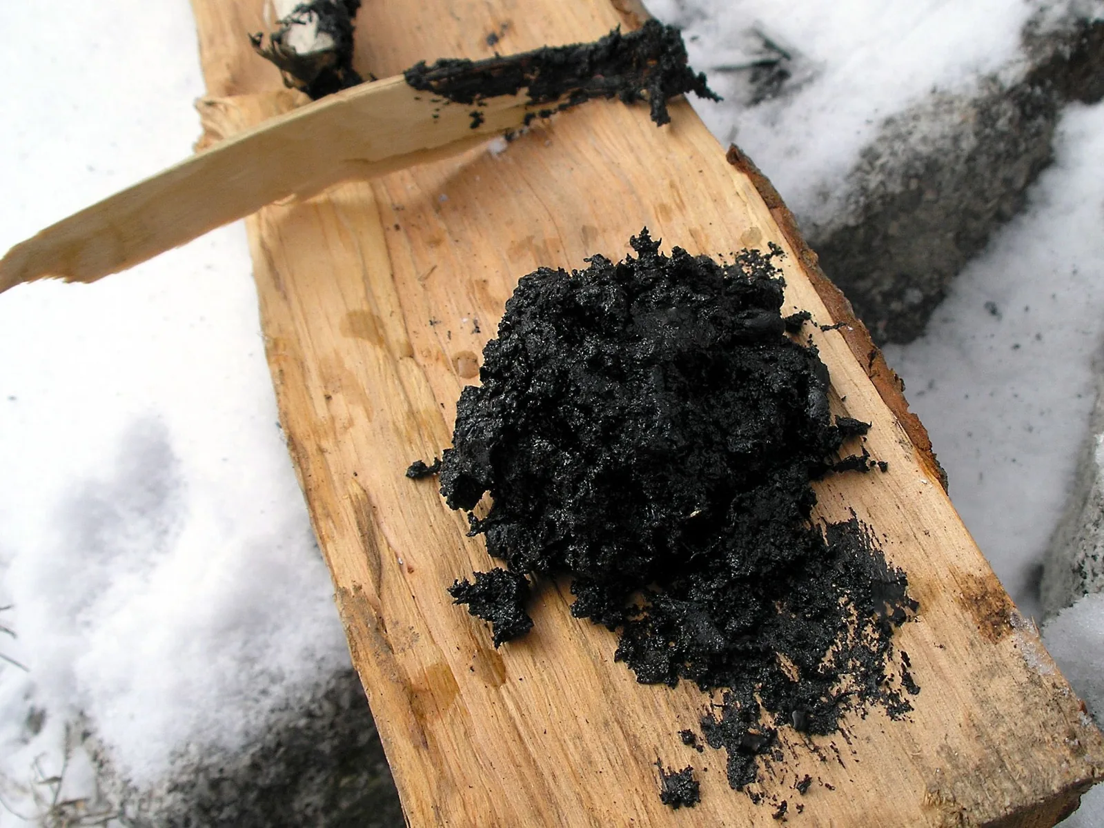 Ein Häufchen schwarzes, krümelig-klebriges Birkenpech liegt auf einem hellen Holzscheit im Schnee; daneben ein Holzspan mit Pechresten.