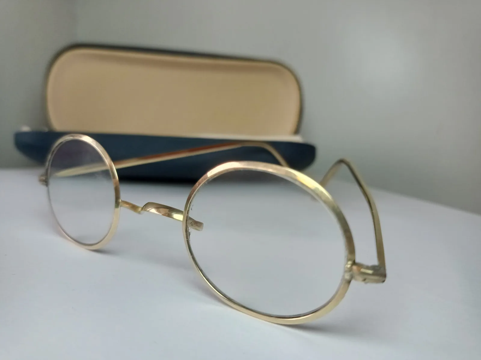Eine Brille mit runden Gläsern und dünnem, goldfarbenem Metallgestell liegt vor einem geöffneten Brillenetui.