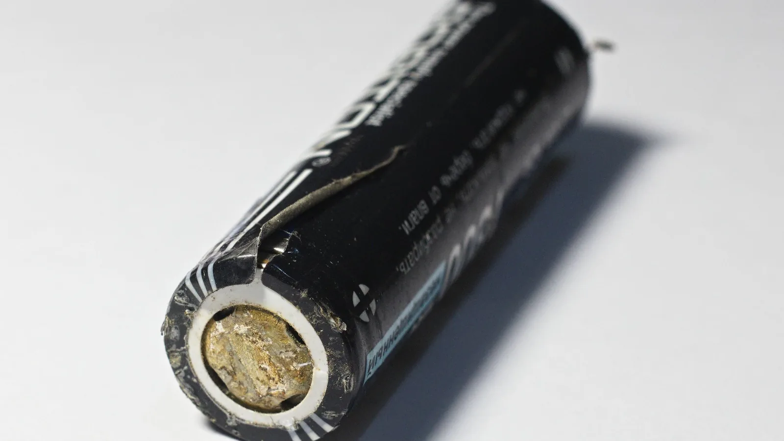 Eine ausgediente zylindrische Lithium-Ionen-Rundzelle liegt auf hellem Grund; die Kunststoffhülle ist eingerissen, der Pluspol ist mit gelblich-grauen Ablagerungen verkrustet.