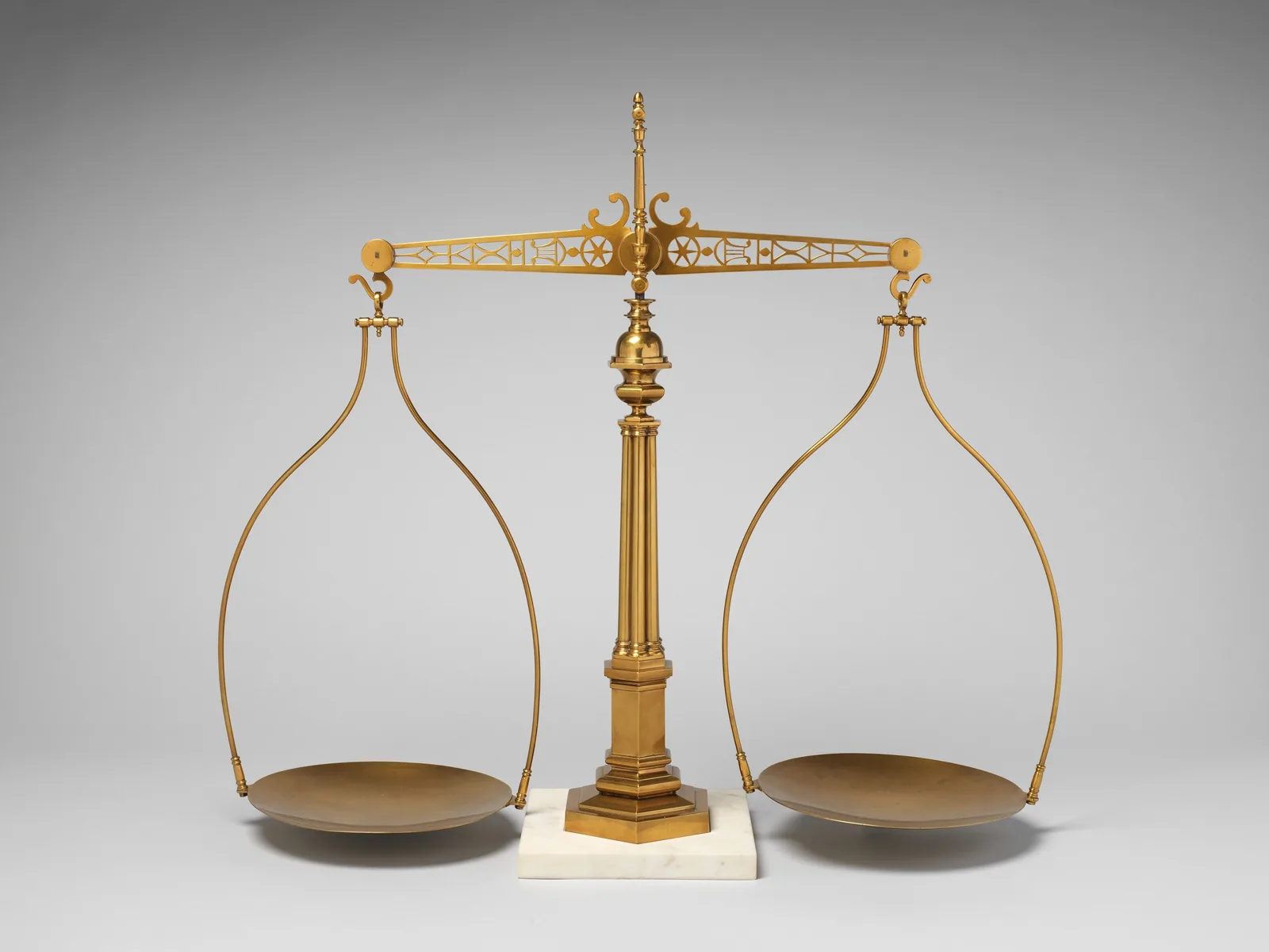 Eine alte Balkenwaage aus Messing: Ein verzierter Balken ruht mittig auf einer Säule, an beiden Enden hängen an Bügeln flache Schalen.