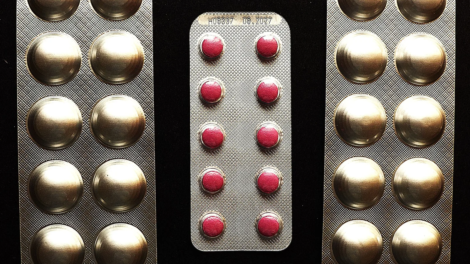 Drei Blisterstreifen auf schwarzem Grund: in der Mitte ein kurzer Streifen mit roten Tabletten, links und rechts längere Streifen mit metallisch glänzenden Mulden.