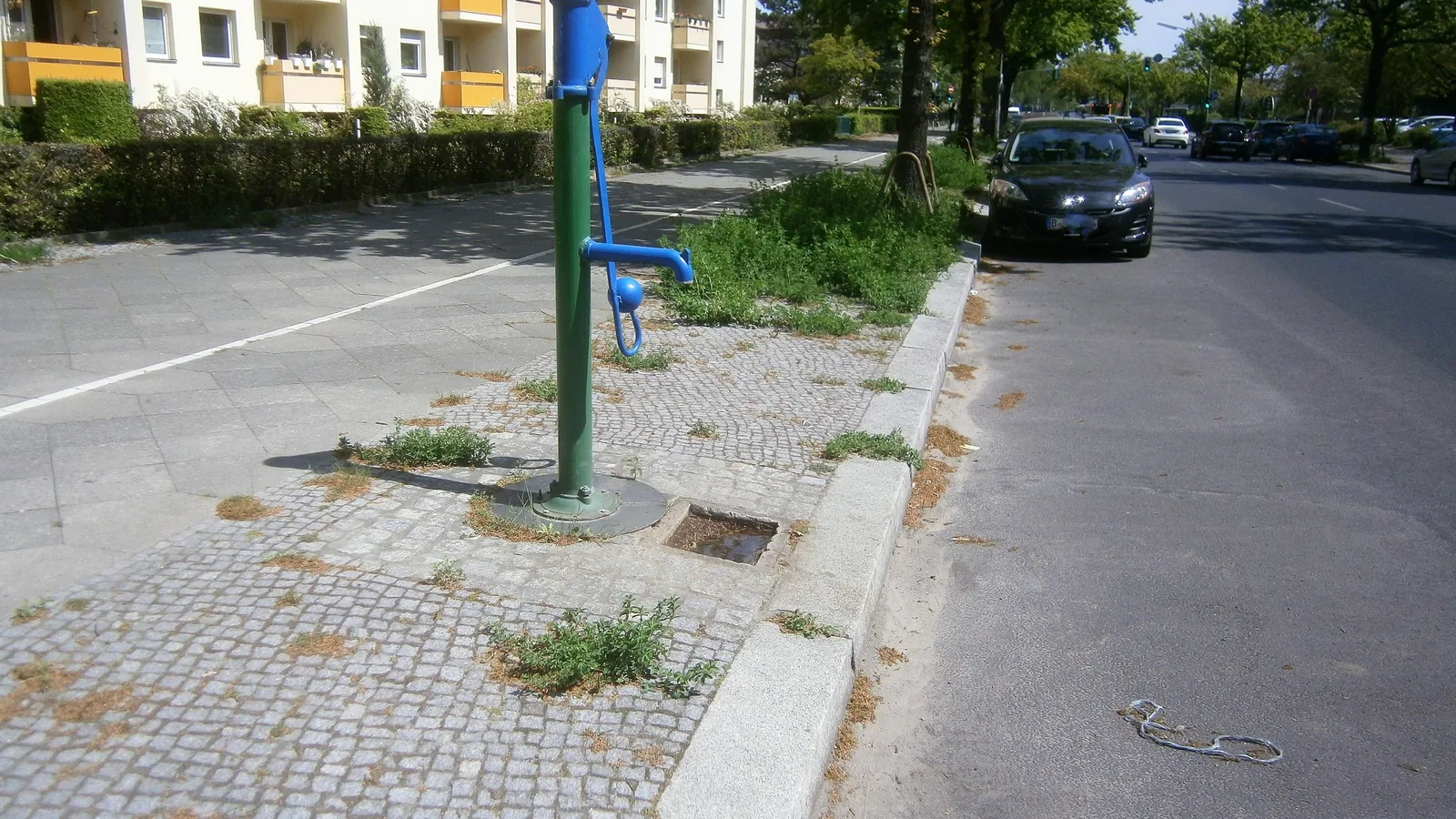 Eine blau und grün lackierte Schwengelpumpe steht auf dem Gehweg einer Straße in Berlin, dahinter Wohnblocks mit Balkonen.
