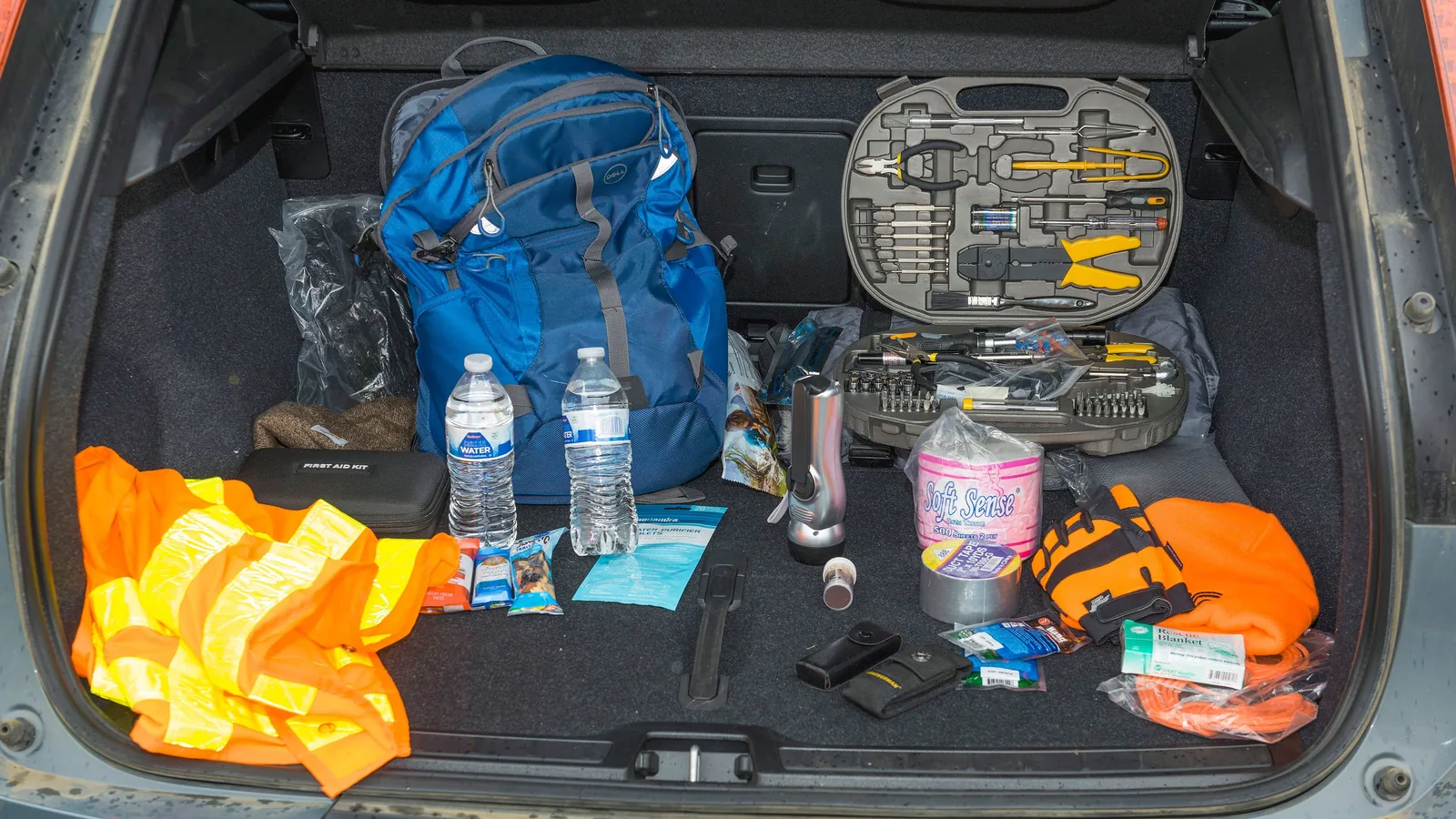 Auf einer Fläche liegen typische Dinge für ein Auto-Notfallset, darunter Verbandmaterial, Wasser, Taschenlampe, warme Kleidung und Werkzeug.