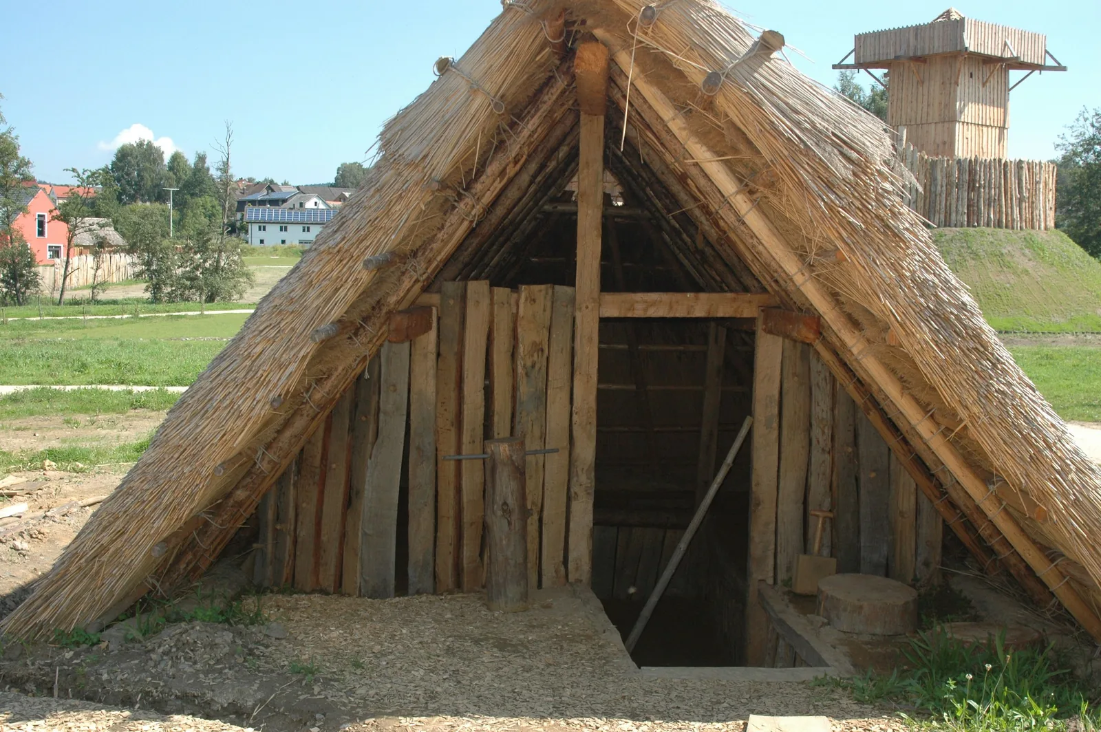 Rekonstruiertes frühmittelalterliches Grubenhaus: ein bis fast zum Boden herabgezogenes, mit Halmen gedecktes Satteldach über einer Giebelwand aus Brettern; der Eingang führt hinab in eine Grube.
