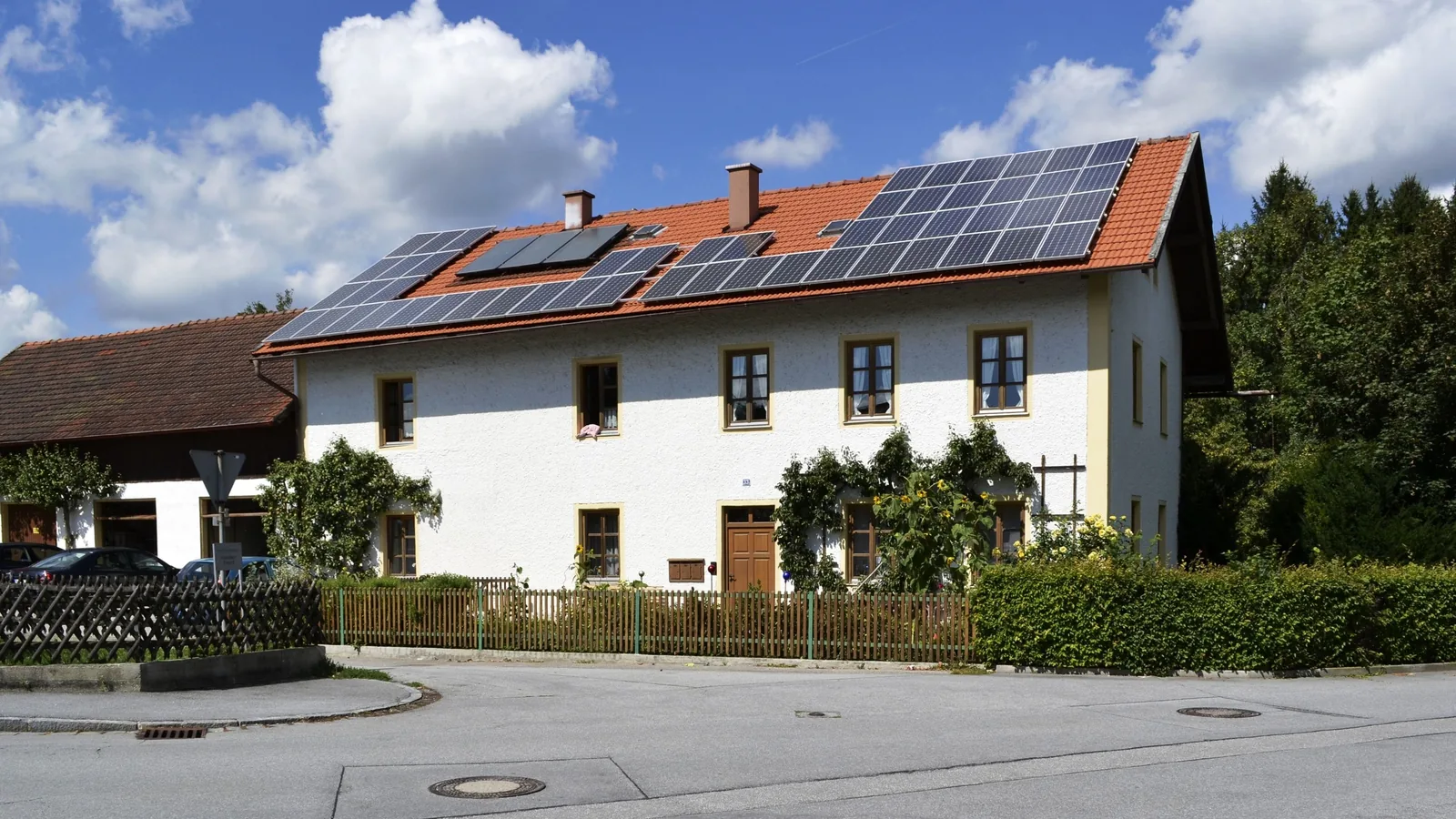 Ein weiß verputztes Wohnhaus mit rotem Ziegeldach, auf dem Photovoltaikmodule montiert sind, an einer Straße in Niederbayern.