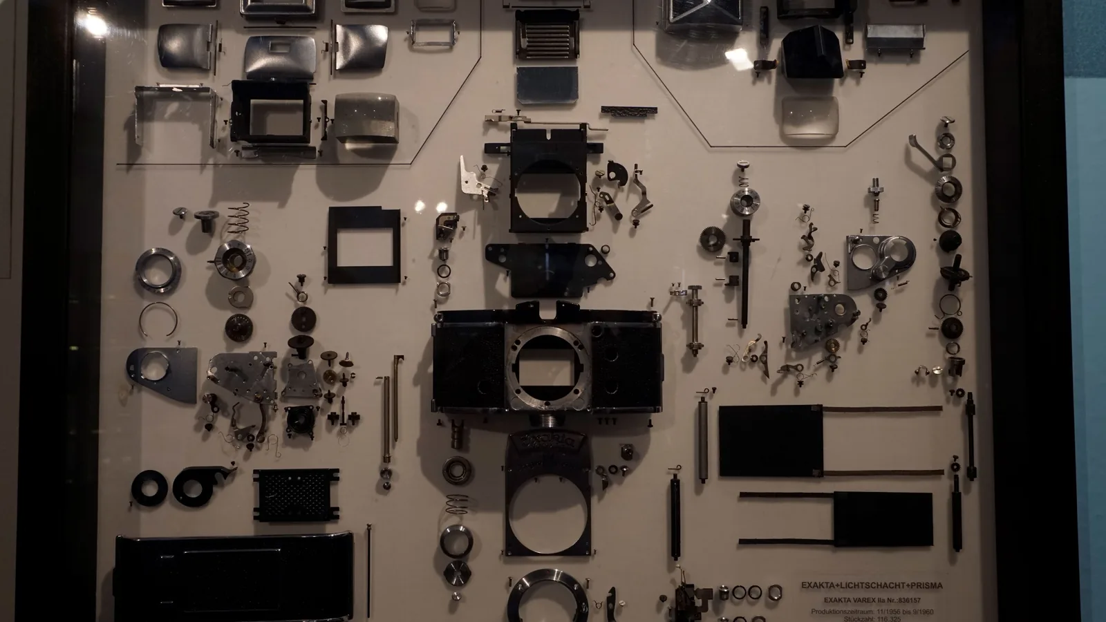 Die zahlreichen Einzelteile einer zerlegten Spiegelreflexkamera liegen geordnet auf einer hellen Tafel in einer Museumsvitrine: Gehäuse, Federn, Zahnräder, Schrauben.