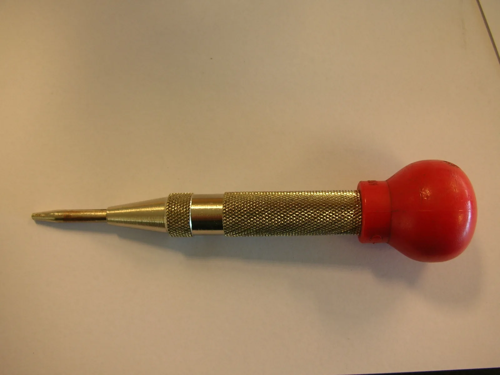 Ein Federkörner auf hellem Grund: ein Metallstift mit feiner Spitze, gerändeltem Griffstück und rotem Knauf.