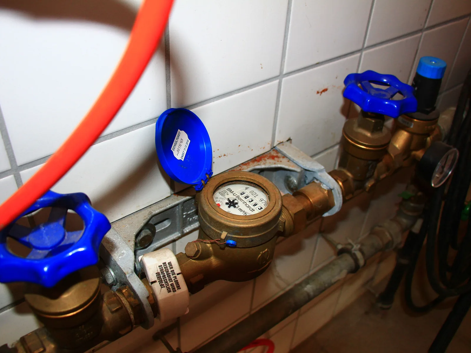 Ein Hauswasserzähler an einer gefliesten Wand, links und rechts davon Absperrventile mit blauen Handrädern.