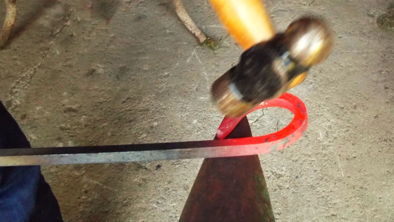 Ein rot glühender Rundstab wird auf dem Horn eines Ambosses mit dem Hammer gebogen; der Hammer ist in Bewegung.