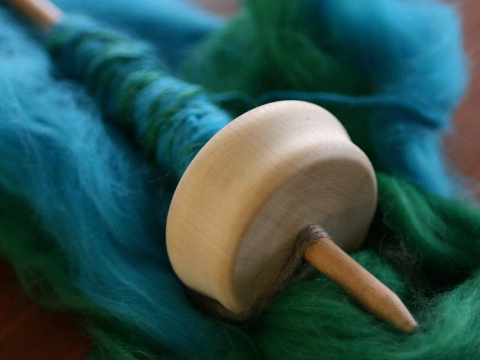 Nahaufnahme einer hölzernen Handspindel mit scheibenförmigem Wirtel auf einem Knäuel blaugrüner Wolle.