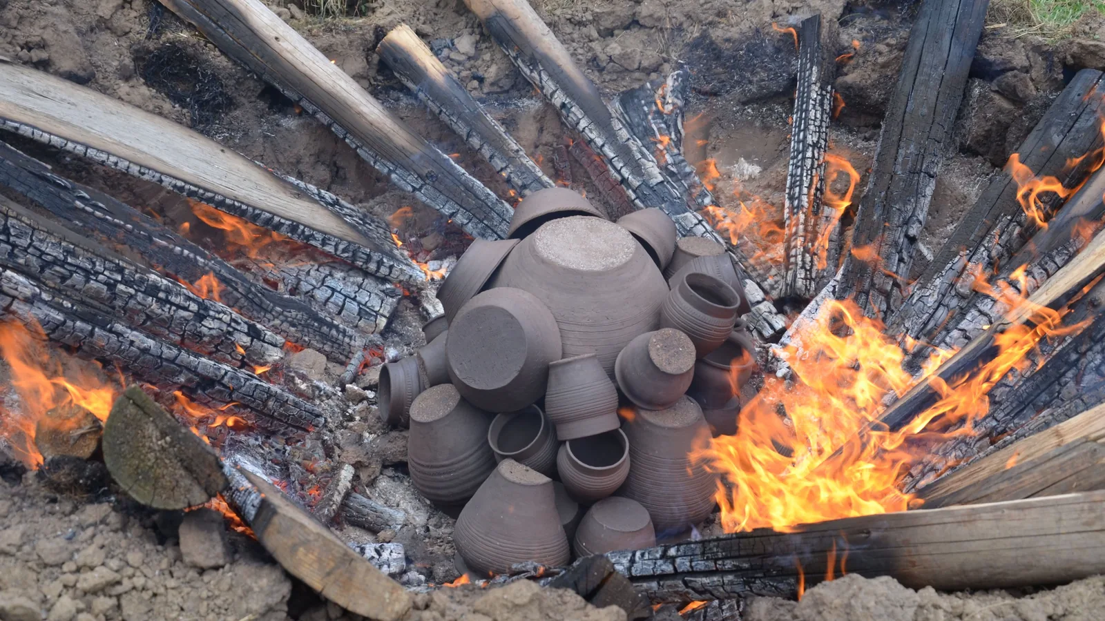 Feldbrand im Freien: Mehrere dunkle, handgeformte Tongefäße stehen dicht beieinander, ringsum sind brennende Holzscheite angelehnt, Flammen schlagen zwischen den Scheiten hoch.