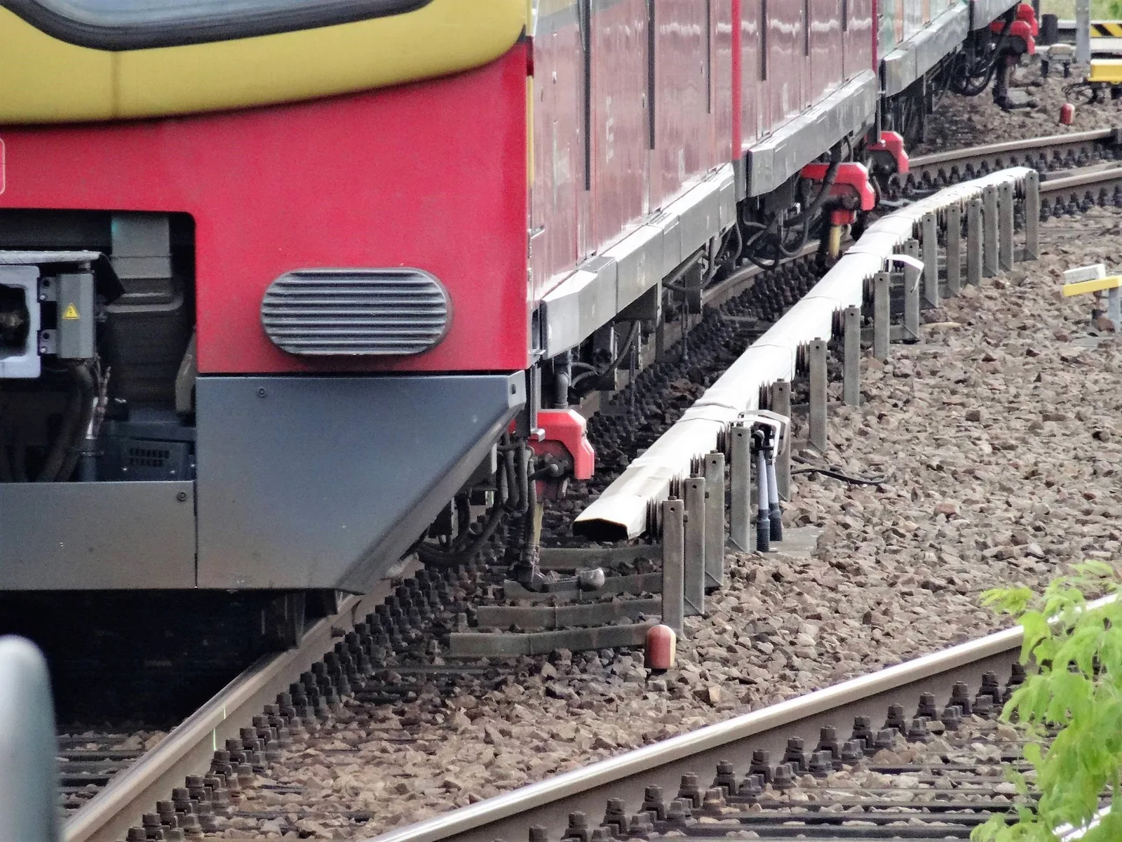Neben dem Gleis verläuft eine abgedeckte Stromschiene; darüber fährt ein rot-gelber S-Bahn-Wagen, dessen Stromabnehmer die Schiene von unten berührt.