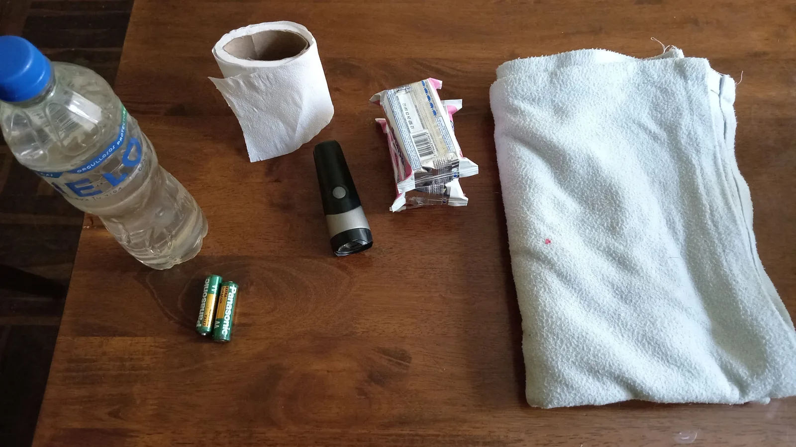 Auf einem Holztisch liegen einfache Haushaltsgegenstände für Notfälle, darunter Wasser, Taschenlampe, Batterien, Handtuch, Toilettenpapier und Lebensmittel.