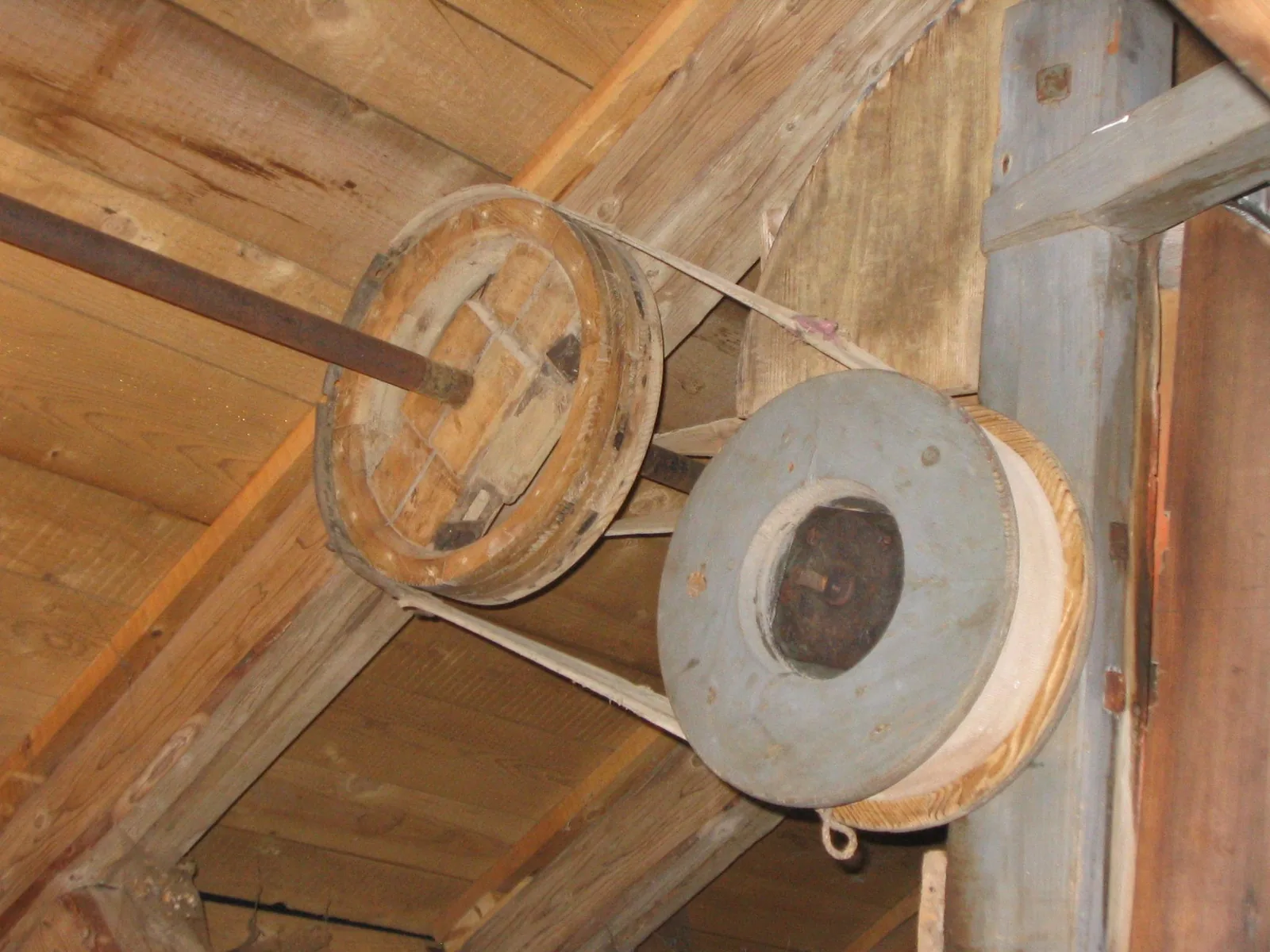 Zwei hölzerne Riemenscheiben an Wellen unter einer Holzdecke, verbunden durch einen Flachriemen.