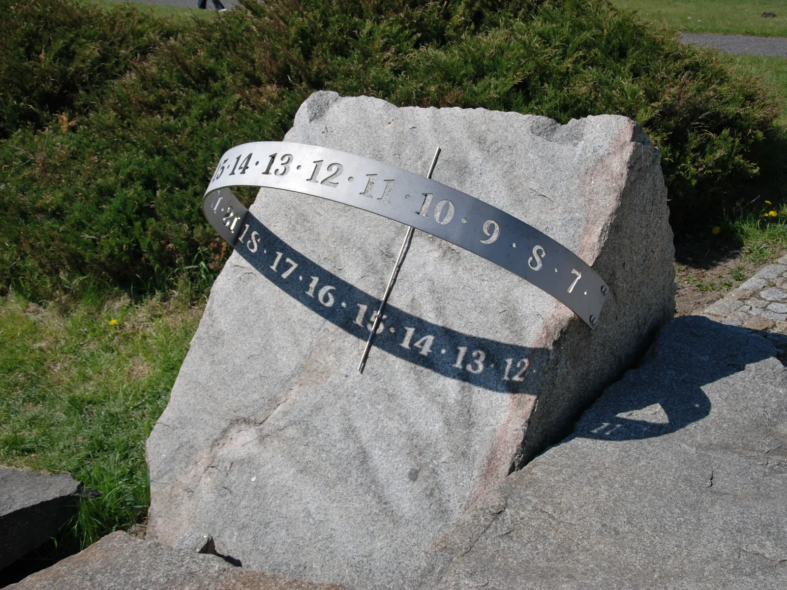 Eine Äquatorialsonnenuhr auf einem Granitblock: Ein gebogenes Metallband mit Stundenzahlen umschließt schräg einen Stab, dessen Schatten auf die Zahlen fällt.